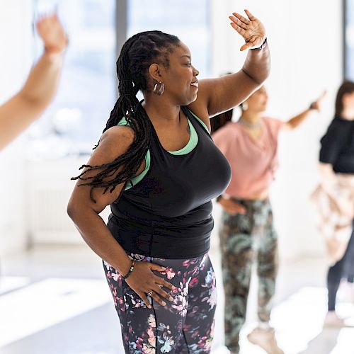 A group of people in a dance class, wearing activewear, engaged in synchronized movement with sunlight streaming through large windows.
