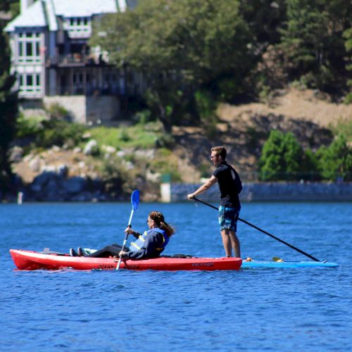 A person is paddleboarding next to someone kayaking on a lake, with houses and trees in the background.