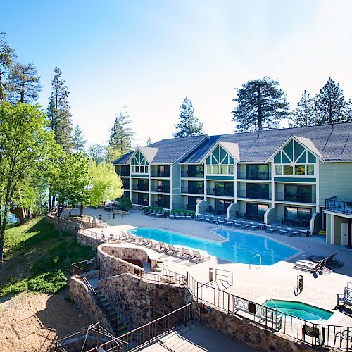 A lakeside resort features a pool, multiple-story glass-fronted buildings, and lush pine trees under a clear blue sky.