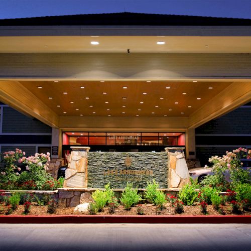 The image shows the entrance of a building with stone pillars, a well-lit ceiling, and landscaped greenery at dusk.