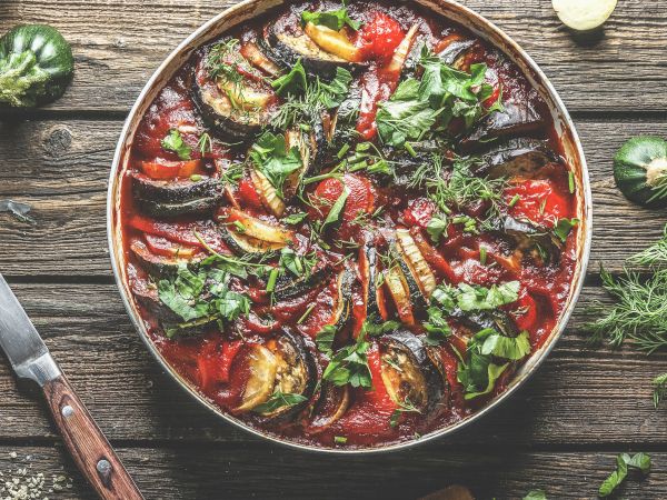 A colorful dish with sliced vegetables, possibly ratatouille, garnished with herbs on a wooden table, surrounded by a knife and more herbs.