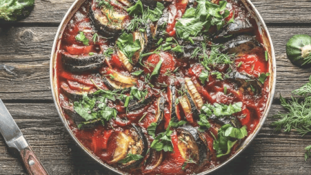 This image shows a dish of ratatouille with sliced vegetables and fresh herbs on a wooden table.