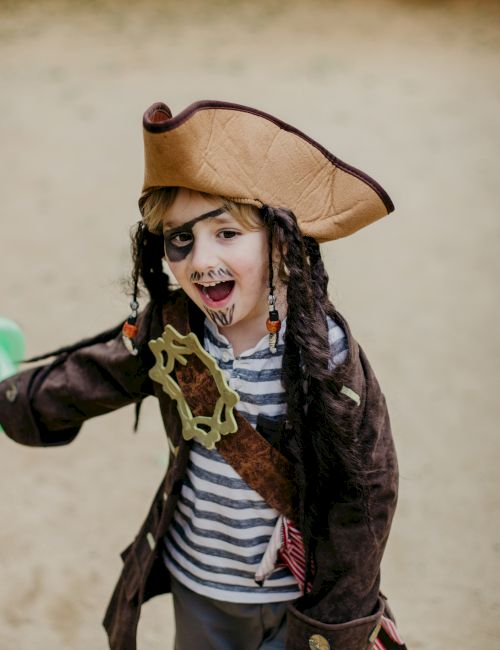 A child dressed as a pirate, with a hat and striped shirt, holding a green toy sword, smiling and playing on a sandy surface.