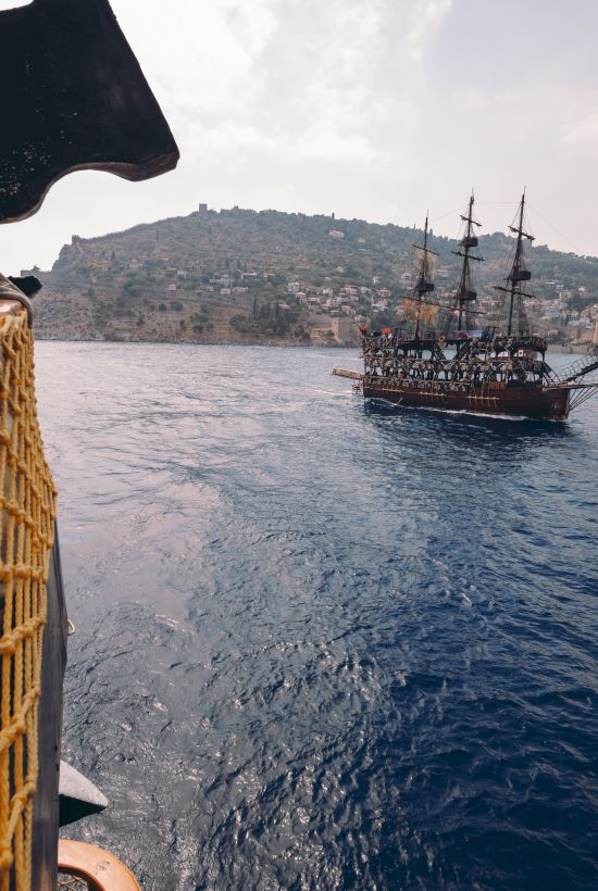 A pirate-style ship and a smaller boat on the water, viewed from another vessel, with hills and buildings in the background under a cloudy sky.