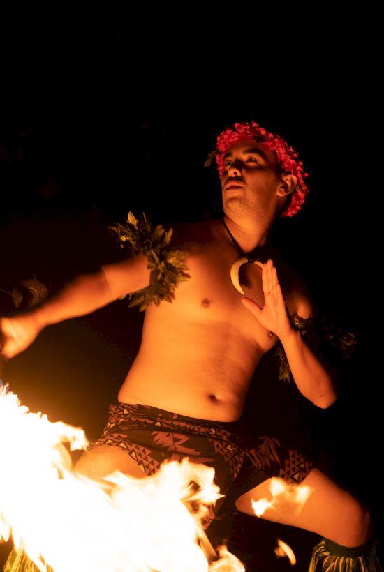 A person is performing a fire dance at night, wearing traditional attire and a floral headpiece, surrounded by flames.