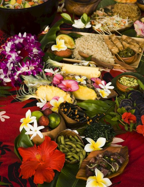 A vibrant tropical spread featuring assorted fruits, flowers, and traditional dishes arranged on a red cloth with decorative elements.