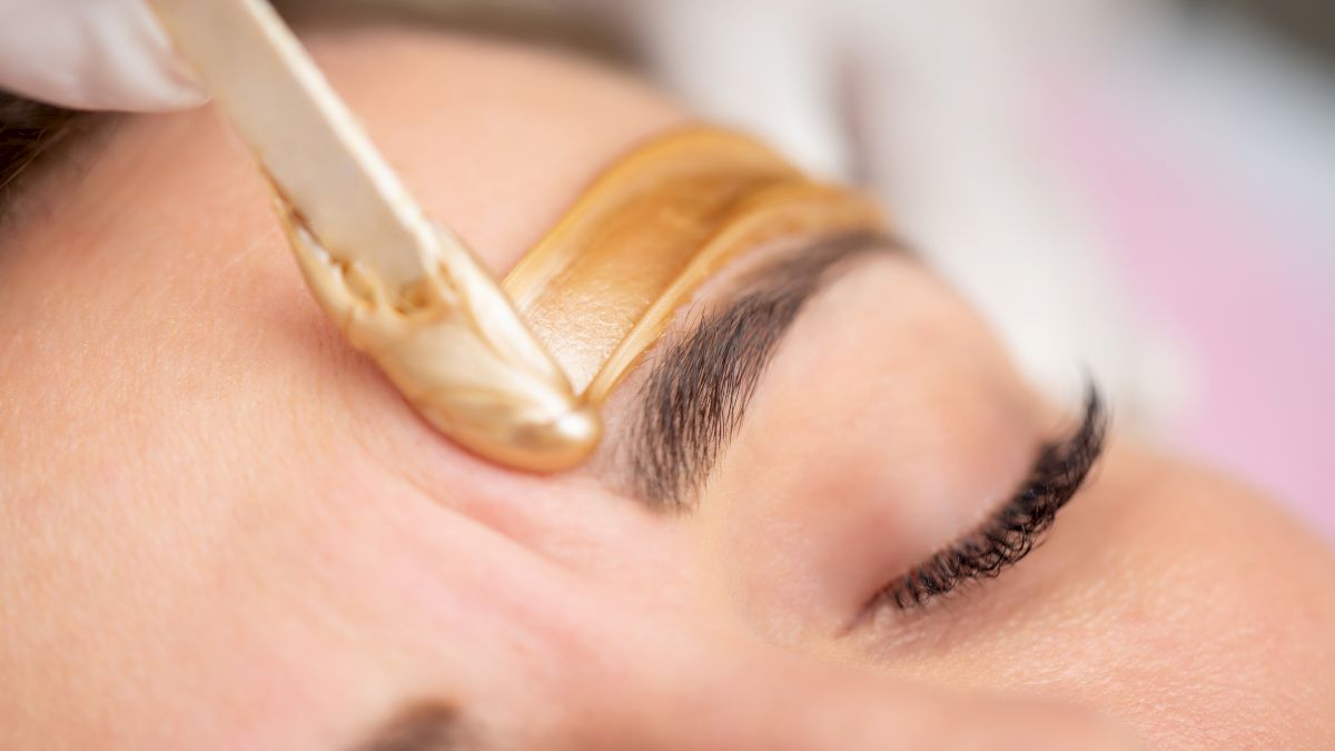 A close-up of a person receiving eyebrow waxing, with a spatula applying wax above the eyebrow.