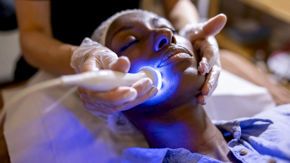 A person in a spa receives a facial treatment with a glowing blue light device, while a practitioner gently supports their head.