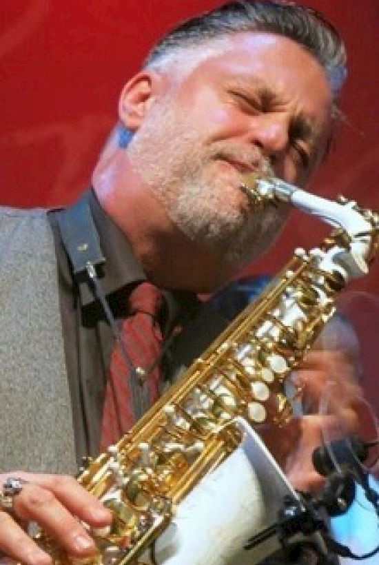 A man in a suit passionately plays the saxophone on stage against a red background.