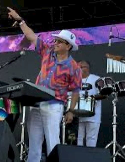 A musician in a colorful shirt and hat performs on stage with a keyboard and drummer.