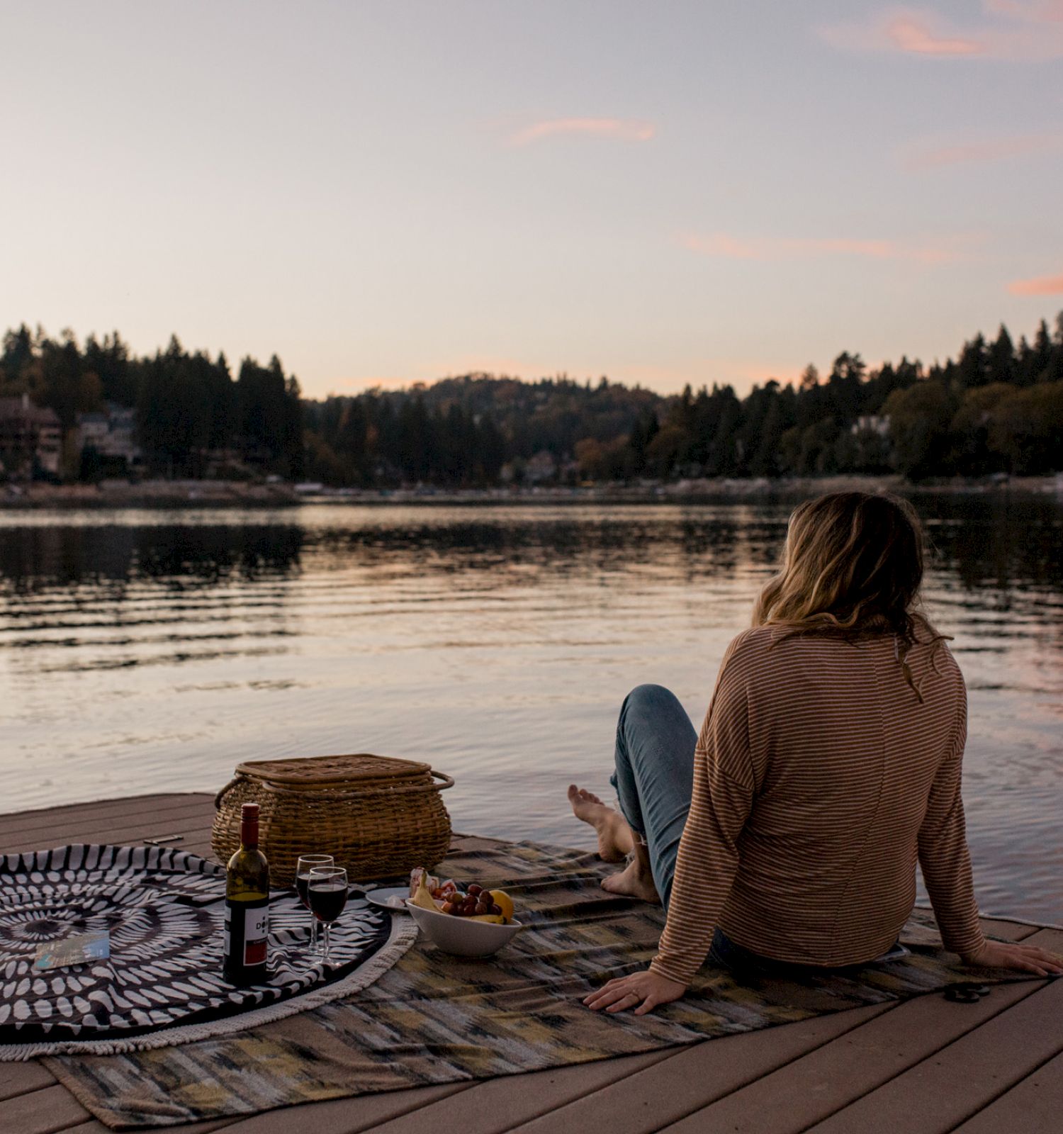 A person sits on a dock by a lake, with a picnic setup, during sunset.