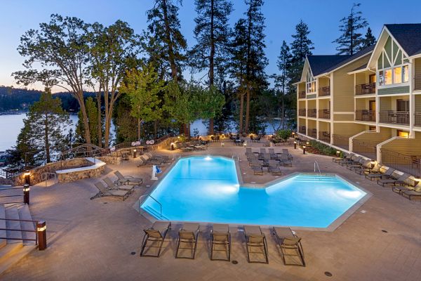 A hotel pool area at dusk with lounge chairs, surrounded by trees. The building is adjacent to a large lake, creating a serene atmosphere.