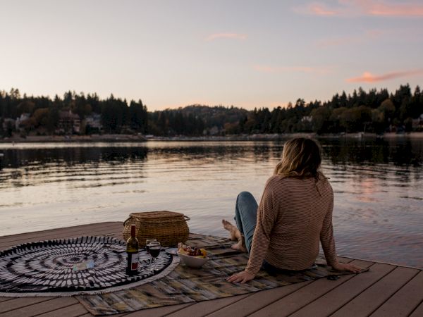 A person sits on a dock by a lake with a picnic setup of a basket and wine at sunset.