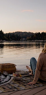 A person sits on a dock overlooking a serene lake, with a picnic setup beside them during sunset.