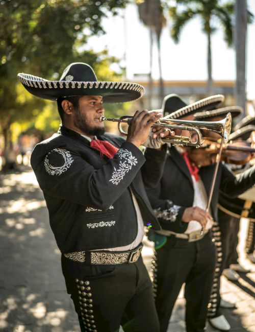A mariachi musician in traditional attire plays a trumpet outdoors, with another musician in the background.