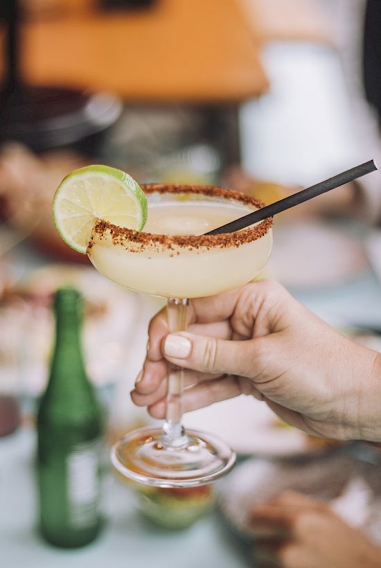 A person is holding a margarita with a lime slice and a black straw at a table with plates, a beer bottle, and people in the background.