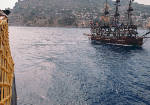 A sailing ship on the sea with hills in the background, viewed from another vessel with a rope railing on the side.