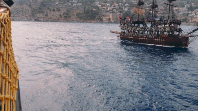 A sailing ship on the sea with hills in the background, viewed from another vessel with a rope railing on the side.
