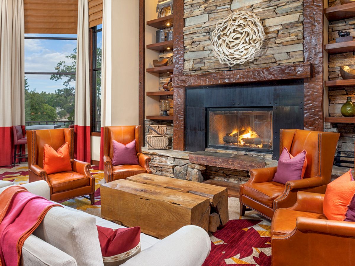 A cozy living room with a stone fireplace, leather chairs, colorful pillows, and a wooden coffee table. Shelves hold decor items around.