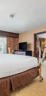 The image shows a hotel room with a bed, TV, and a seating area. There are curtains on the windows and a chair beside a table.